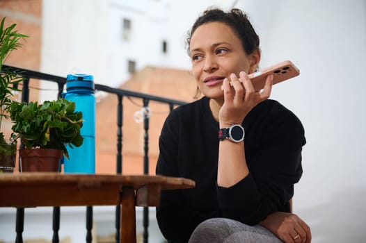 A woman relaxes on a sunny balcony, smiling while talking on her smartphone. She is dressed casually and is surrounded by plants and a water bottle, capturing a peaceful moment.
