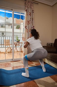 A woman performs a squat exercise indoors using a resistance band on a yoga mat. The bright living room setting, with natural light and vibrant details, emphasizes home workout commitment.