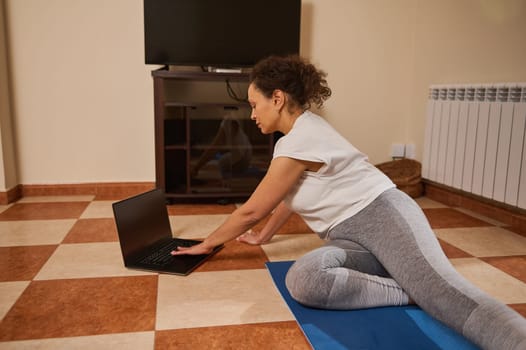 A woman is stretching on a yoga mat while following an online yoga class at home. She uses a laptop for her virtual practice, combining fitness with convenience and relaxation in her living space.