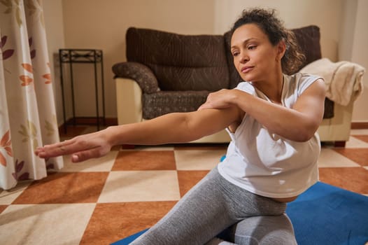 A woman practices stretching exercises at home, sitting on a yoga mat. The warm, serene indoor setting emphasizes relaxation and wellness during her fitness routine.