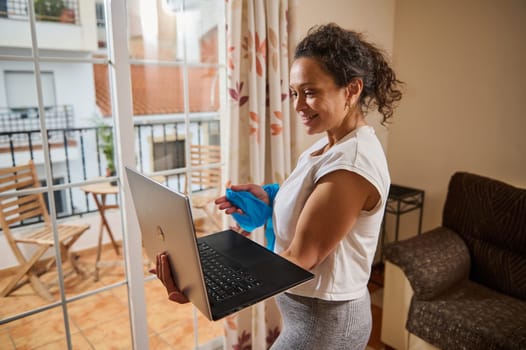 A happy woman engages in a virtual fitness class at home, holding a laptop while smiling. She stands in a bright living room, creating a positive and active atmosphere.