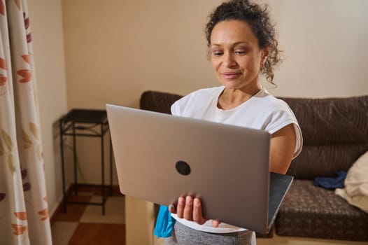 A woman stands in a cozy living room holding a laptop, engaging in remote work or virtual communication. She appears focused and comfortable in her casual attire.