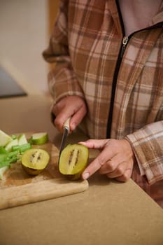 A person wearing a plaid jacket slices a kiwi fruit on a wooden cutting board. The scene evokes a cozy, homey atmosphere, perfect for healthy living and culinary inspiration.
