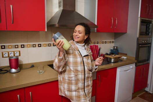 A cheerful woman in a cozy plaid jacket holds a nutritious green smoothie in a bright, modern kitchen with red cabinets. Emphasizing healthy lifestyle and joyful moments at home.
