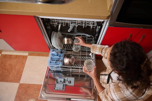 A woman is seen loading glasses into an open dishwasher in a modern kitchen with red cabinetry. The setting is casual, reflecting daily routine and efficient home management.