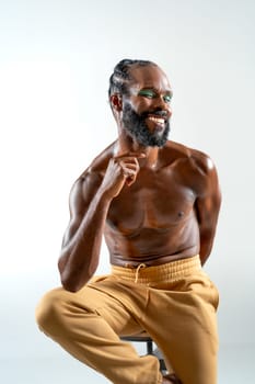 Happy African American shirtless gay model sitting on stool over white background. Portrait of smiling homosexual man is posing in plain studio.