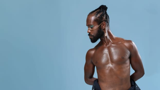 African American shirtless gay model with hands behind back looking down against blue background. Serious homosexual man standing in plain studio.