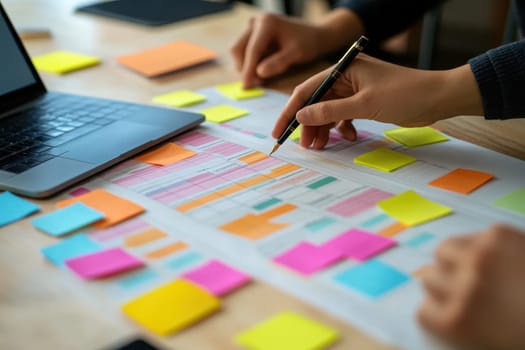 Two hands engaged in planning with colorful sticky notes on a project overview layout beside a laptop. Ideal for teamwork and strategy visuals.