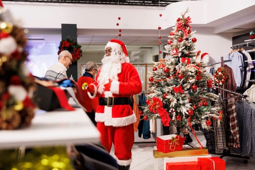 Employee Santa Claus in Christmas themed store, jingling xmas bells to attract customers in shopping center. Retail assistant dressed in festive costume greeting clients in decorated fashion boutique