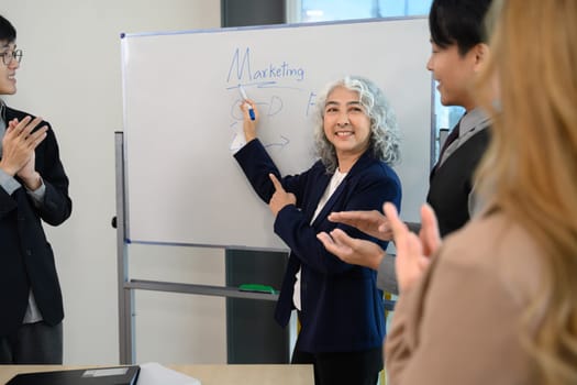 Mature corporate leader presenting marketing strategy on whiteboard and receiving applause from her team.