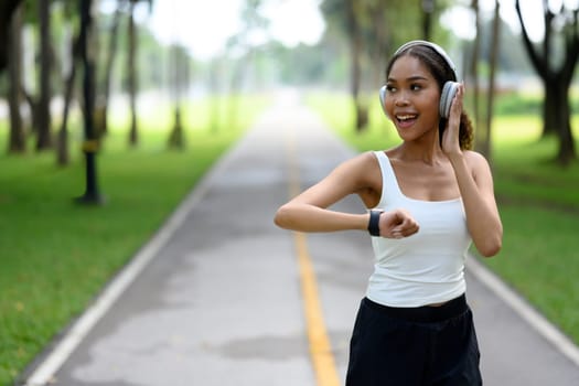 Cheerful young sportswoman monitoring her progress on a smartwatch while jogging through a peaceful park.