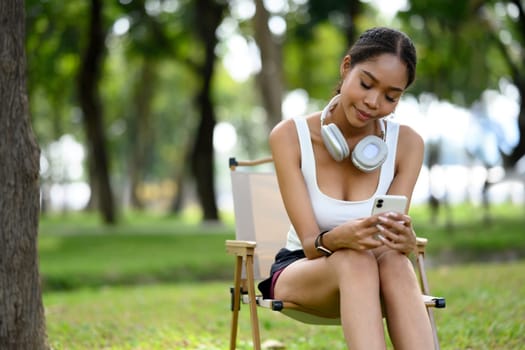 Pretty African American woman using mobile phone a portable chair in the park taking a break after her workout.