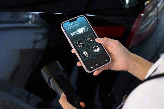 Woman using EV application on smartphone to check charging status while plugged in the EV charging connector.