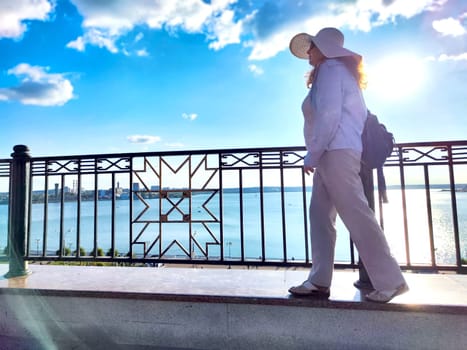 A woman strolls near the water's edge, sporting a hat and light clothing under clear blue sky filled with sunlight