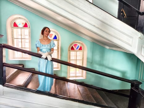 A girl in a vintage blue dress stands elegantly on a staircase in an ancient castle, surrounded by stunning stained glass windows.