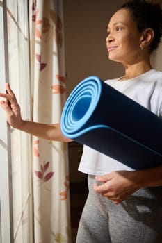 A woman stands in a sunlit room holding a yoga mat, looking thoughtfully out the window. Her demeanor suggests calm and readiness for exercise, embracing mindfulness and tranquility.