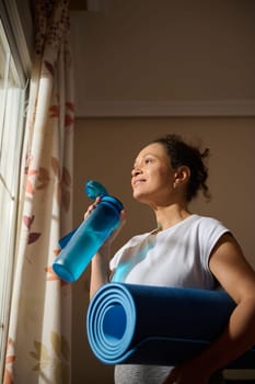 A woman stands by the window holding a yoga mat and a water bottle, looking outside with a hopeful expression. Her relaxed demeanor suggests preparation for a peaceful yoga session.