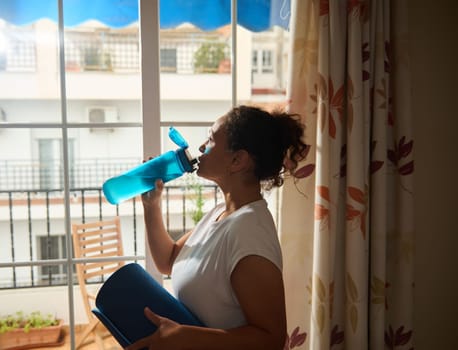 A woman rehydrates with a large water bottle after a yoga session at home, holding a blue yoga mat. She stands by a sunlit window, looking relaxed and refreshed. Ideal for healthy living themes.