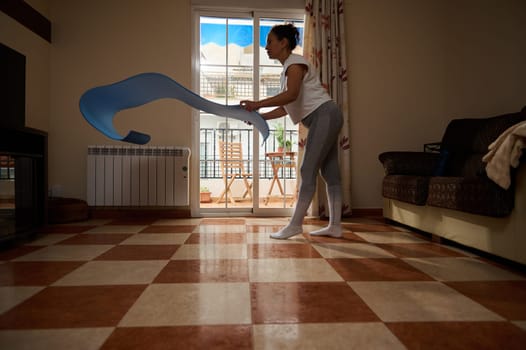 A woman engages in a stretching routine using a blue fitness band in her living room. Natural light fills the space, creating a calming environment for a focused workout session.