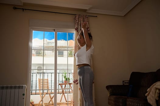 A woman performs a yoga stretch indoors with sunlight streaming through a window. The serene atmosphere enhances focus and mindfulness, reflecting a peaceful start to the day.