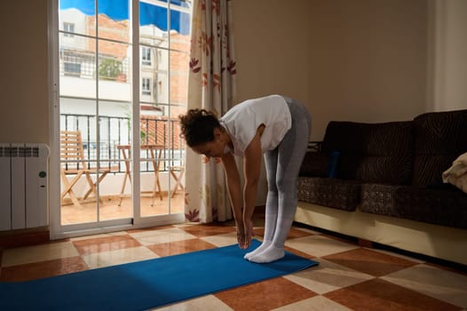 A woman stretches on a blue yoga mat in a cozy living room with soft sunlight streaming through the window. Emphasizing flexibility, relaxation, and a peaceful home atmosphere.
