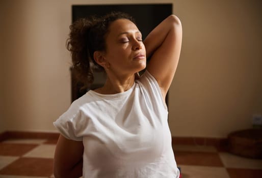 A woman enjoying a moment of mindfulness while stretching in her home yoga session. The soft light highlights her calm and peaceful expression, capturing a sense of tranquility and focus.