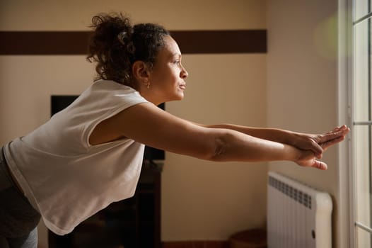 A woman is practicing yoga indoors, stretching forward with focus and calmness. The natural light highlights her peaceful expression, embodying balance and mindfulness.