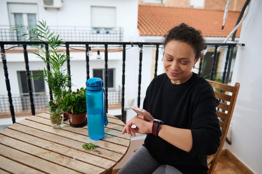 A woman sitting on a balcony checks her smartwatch, surrounded by green plants and a blue water bottle. The scene is calm and serene, highlighting relaxation and mindfulness.