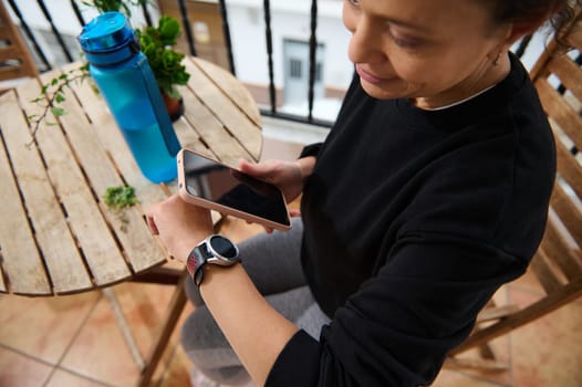 A woman sits on a balcony, checking notifications on her smartwatch while holding a smartphone. A water bottle and plant are on a nearby table, reflecting a relaxed outdoor setting.
