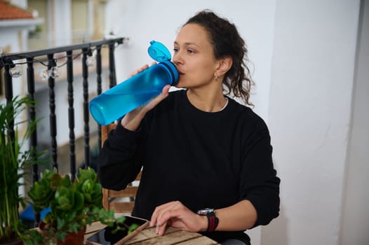 A woman sips water from a blue bottle on her balcony, surrounded by potted plants, capturing a serene moment of relaxation and hydration.