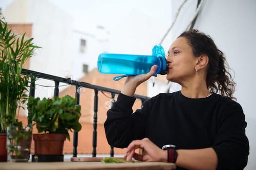 A woman enjoys a refreshing drink from a water bottle while sitting on a balcony. The scene is surrounded by green plants, creating a calm and serene environment.