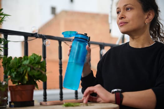 A woman sits on a balcony, holding a blue water bottle, surrounded by plants. She appears relaxed in the sunlight, symbolizing hydration and tranquility in an outdoor setting.