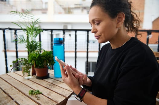 A woman sits on a balcony table texting on her smartphone. Surrounded by greenery and plants, she enjoys a tranquil moment in an urban setting, with a water bottle in the background.