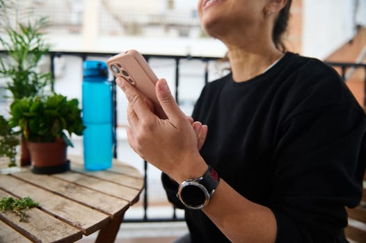 A woman relaxes on a balcony, enjoying a moment with her smartphone. The scene is cozy, featuring plants and a water bottle, conveying tranquility and leisure.