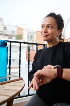 A thoughtful woman sits on a balcony checking her smartwatch while staying hydrated with a water bottle beside her. The image conveys a sense of mindfulness and a healthy lifestyle.