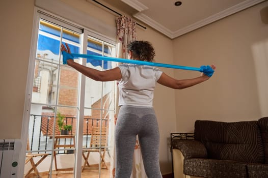 A woman stands by a large window performing a resistance band exercise. She stretches her arms outward, focusing on fitness and well-being in the comfort of her living room.