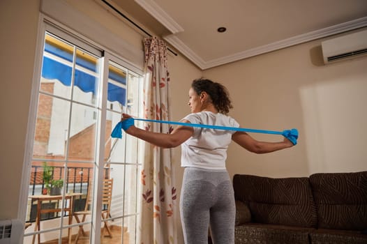 A woman performs a resistance band exercise in her living room, emphasizing fitness and home workout routines. She stretches her arms with focused determination, promoting a healthy lifestyle.
