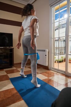 A young girl engages in a fitness routine indoors, using a blue yoga mat and resistance band. The sunlight streaming through the window creates a peaceful workout environment.
