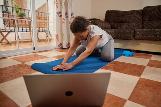 A woman doing a yoga stretch on a mat in her living room, following an online class. The setting is cozy with a sofa nearby and patio view. It promotes wellness and home exercise.
