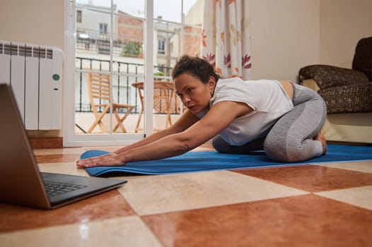 A woman performs a yoga stretch on a blue mat at home, following an online class on her laptop. The comfortable indoor setting enhances her focused and relaxed demeanor.