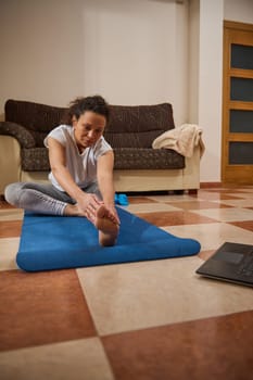 A woman performs yoga on a mat in her living room while following an online session. She is focused and committed to her fitness routine, demonstrating dedication and serenity.