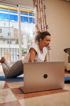 A woman concentrates on her yoga routine at home, following along with an online class on her laptop. The bright room and focused expression indicate a dedicated fitness session.