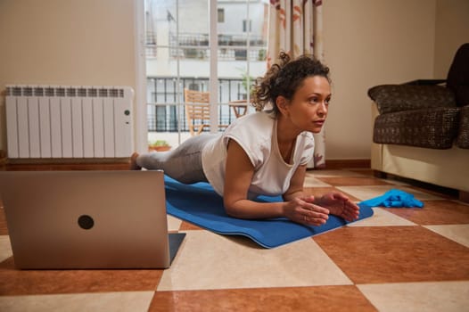 Focused woman in athletic wear doing a yoga plank on a mat at home. She uses a laptop for virtual guidance, balancing fitness and comfort in her living space.