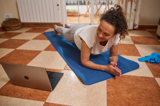 A woman is practicing an online workout at home, doing a plank on a yoga mat. She is following a virtual exercise video on her laptop in a living room setting.