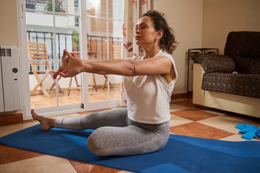 A middle-aged woman practices yoga in her living room, sitting on a mat and stretching her arms. This indoor exercise scene embodies mindfulness and flexibility in home fitness.