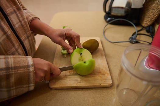 A person carefully slices a fresh green apple on a wooden board in the kitchen. The cozy setting suggests a focus on healthy eating and meal preparation, with a kiwi nearby.