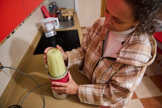 A woman in a cozy kitchen setting prepares a fresh smoothie using a blender. The scene portrays a healthy lifestyle and home cooking, emphasizing the trend of nutritious, homemade beverages.