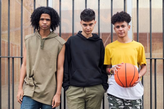 three multiracial male teenagers with basketball standing together on street and looking at camera, concept of friendship and urban lifestyle