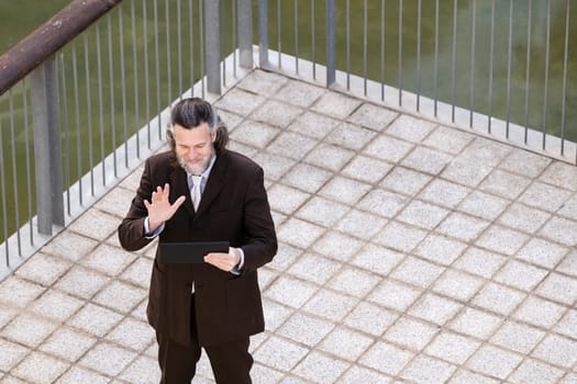 happy middle age male with beard in elegant suit waving hand while greeting partner during video call via digital tablet on terrace, concept of business and mature man lifestyle, copyspace for text
