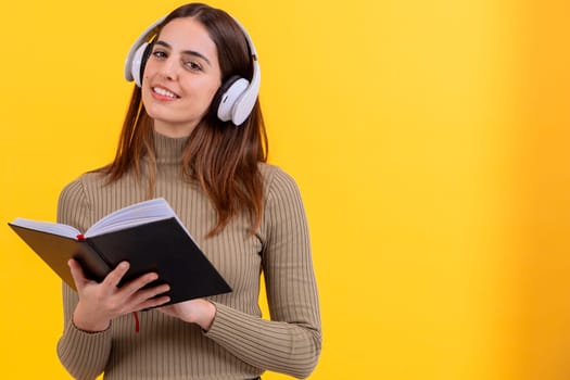 A cheerful young woman wearing white headphones and holding an open book, standing against a vibrant yellow background. Perfect image for learning, audiobooks, or educational themes.copy space